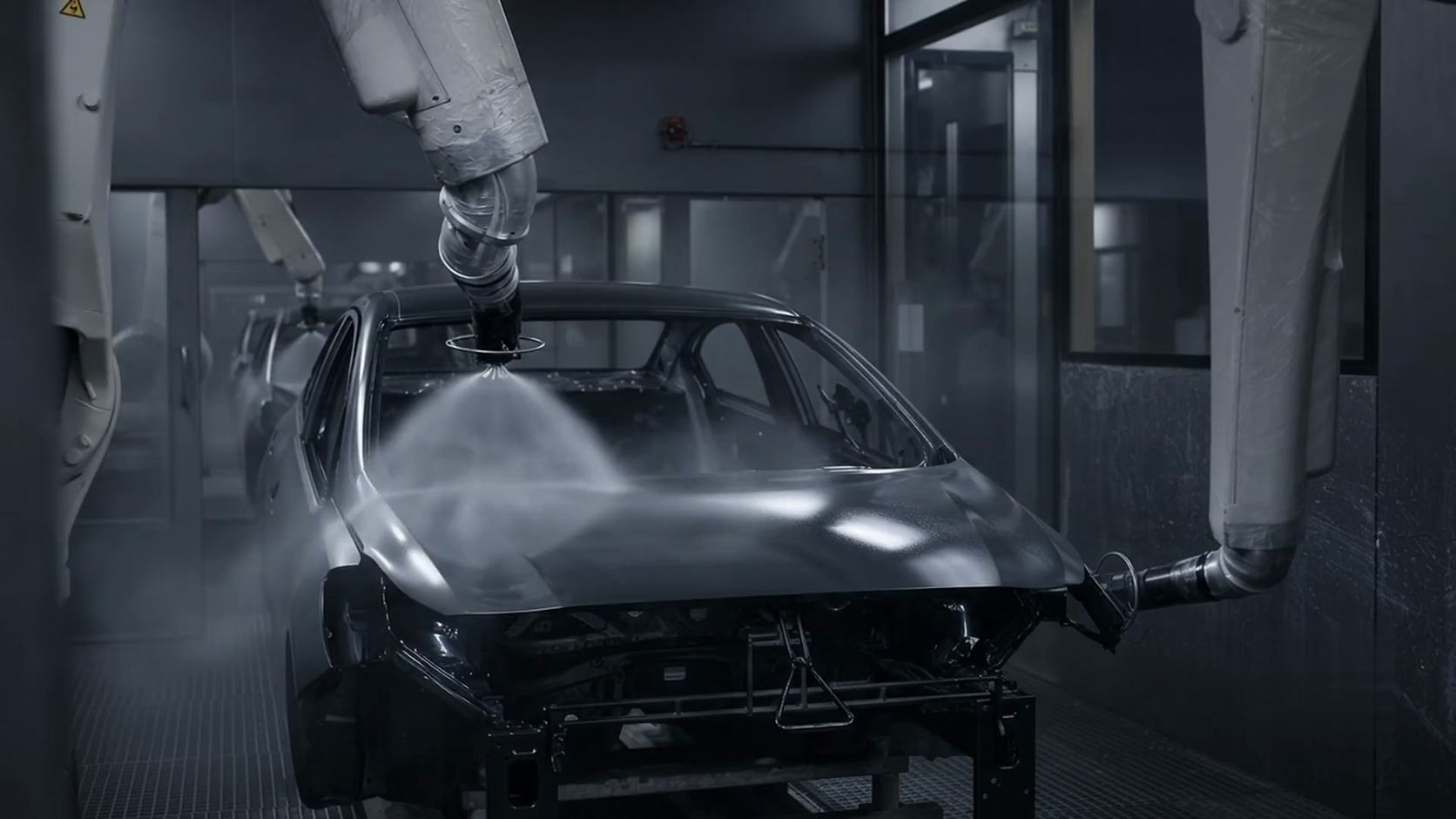 A car body in a real automotive paint booth, an overhead applicator laying down paint while fine mist drifts through the booth light